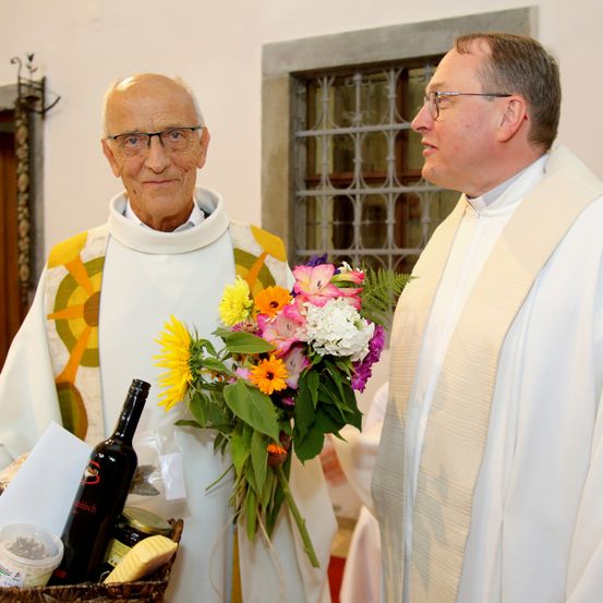 Zwei Priester stehen nebeneinander. Der ältere Priester hält einen Blumenstrauß. Der jüngere Priester schaut den älteren Priester an. Ein Korb mit einer Flasche Wein, Marmelade und Käse steht vor ihnen.