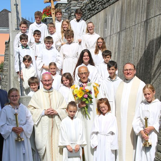 Eine Gruppe von Menschen in weißen Gewändern, möglicherweise Priester, steht auf einer Betonfläche. Sie posieren für ein Foto mit Blumen und Kerzen. Dahinter befindet sich eine Wand mit einem Kreuz und einem Steinpfeiler.