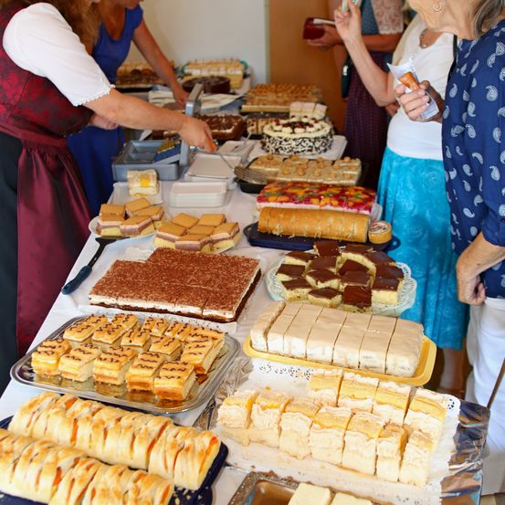 Ein langer Tisch ist mit einer Vielzahl von Desserts beladen, darunter Kuchen, Gebäck und Torten. Mehrere Frauen stehen herum und bewundern das Angebot. Eine Frau hält ein Getränk in der Hand.