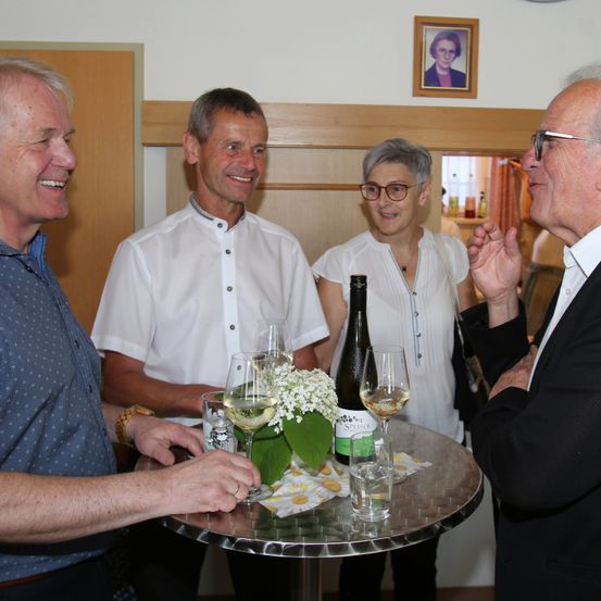 Vier Menschen lächeln und plaudern um einen Tisch mit Weinläsern und einer Flasche Wein. Dahinter hängt ein gerahmtes Bild an der Wand.