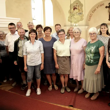 Eine Gruppe von Menschen posiert für ein Foto in einer Kirche. Sie stehen im Halbkreis, lächeln und sind in lässiger Kleidung gekleidet. Hinter ihnen ist eine Wand mit einem Gemälde und einem Kreuz zu sehen.