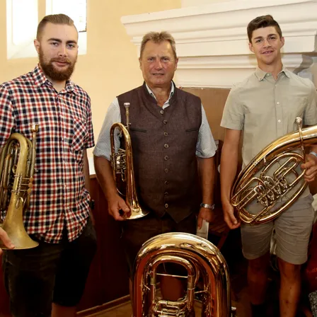 Drei Männer posieren mit Blechblasinstrumenten in einem Raum. Der Mann auf der linken Seite hält eine Trompete, der Mann in der Mitte hält ein Tenorhorn und der Mann auf der rechten Seite hält eine Tuba.
