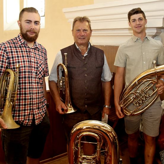 Drei Männer posieren mit Blechblasinstrumenten in einem Raum. Der Mann auf der linken Seite hält eine Trompete, der Mann in der Mitte hält ein Tenorhorn und der Mann auf der rechten Seite hält eine Tuba.