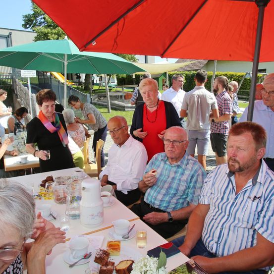 Eine Gruppe älterer Menschen versammelt sich um einen Tisch, möglicherweise bei einer Gemeinschaftsveranstaltung. Einige sitzen, andere stehen. Eine ältere Frau in einem roten Shirt spricht mit einem Mann im weißen Hemd. In der Nähe steht eine Frau in Schwarz und hält ein Glas. Der Tisch ist mit Essen und Getränken gedeckt. Im Hintergrund befinden sich Schirme und ein Gebäude.