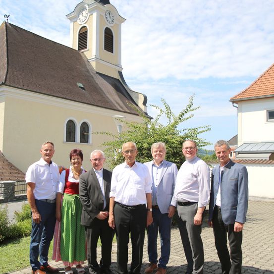 Sieben Menschen stehen vor einer Kirche, ein Mann in Anzug und andere in weißen Hemden. Ein Baum und ein Glockenturm sind im Hintergrund zu sehen.