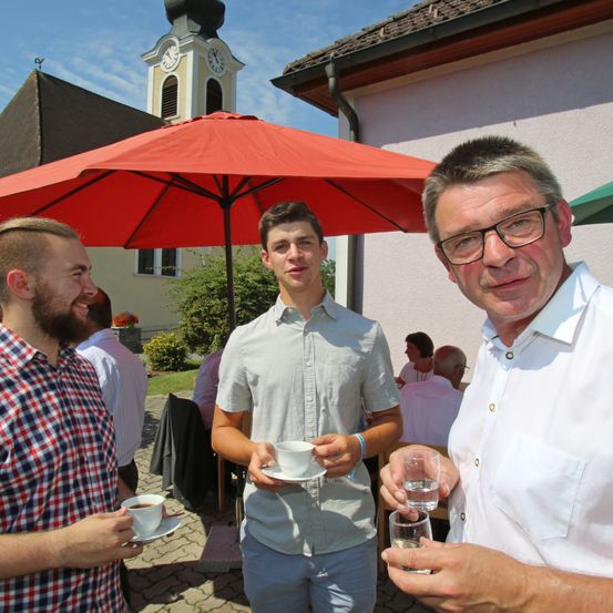 Drei Männer unter einem roten Schirm; einer hält eine Kaffeetasse, der andere ein Wasserglas. Ein Kirchturm im Hintergrund.