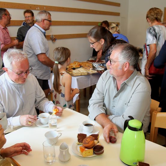 Mehrere Erwachsene und Kinder versammeln sich um einen Tisch mit Essen und Getränken. Zwei ältere Männer unterhalten sich im Sitzen. Eine Frau mit Brille und ein Mädchen mit einem Pferdeschwanz sind bei einem Tisch mit Desserts.