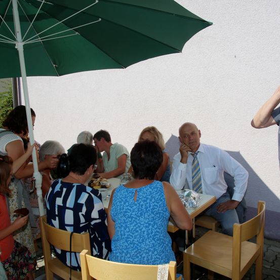 Eine Gruppe von Menschen sitzt unter einem grünen Schirm draußen am Tisch. Ein Mann in weißem Hemd und Krawatte sitzt mit der Hand am Kinn. Eine Frau in blauem Kleid sitzt neben ihm. Eine andere Frau hält eine Kamera.