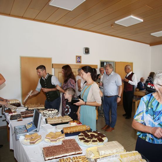 Eine Gruppe von Menschen versammelt sich um einen Tisch voller verschiedener Desserts. Der Tisch ist mit einem weißen Tuch bedeckt. Die Menschen stehen und einige halten Teller. Der Raum hat Holzwände und eine Holzdecke. Es gibt Bilderrahmen an der Wand.