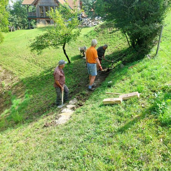 Drei Männer arbeiten auf einem Hügel, einer mit einer Schaufel, ein anderer mit einem Pickel und der dritte mit einer Harke. Im Hintergrund befindet sich ein Haus am Hang.