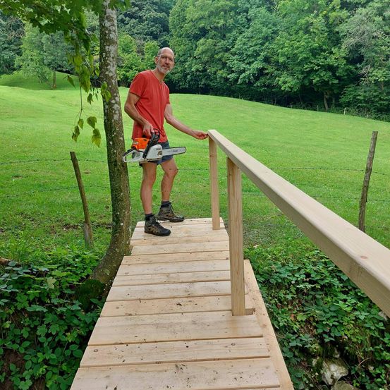 Ein älterer Mann steht auf einer Holzbrücke in einem Grasfeld und hält eine Kettensäge. Er trägt ein rotes T-Shirt und schwarze Shorts. Die Brücke hat ein hölzerne Handgeländer. Im Hintergrund befindet sich ein Stacheldrahtzaun.