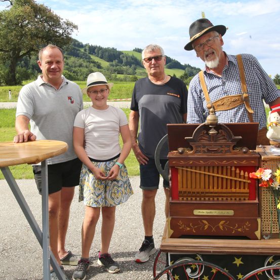 Drei Männer und ein junges Mädchen stehen neben einem alten Musikinstrument, möglicherweise einer Ziehharmonika, in einer malerischen Umgebung im Freien. Sie scheinen für ein Foto zu posieren.