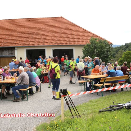 Eine große Gruppe von Menschen versammelt sich um Tische im Freien vor einem Gebäude. Einige sitzen, andere stehen. In der Nähe sind Fahrräder geparkt. Das Gebäude hat ein rotes Dach und ist von Bäumen umgeben.