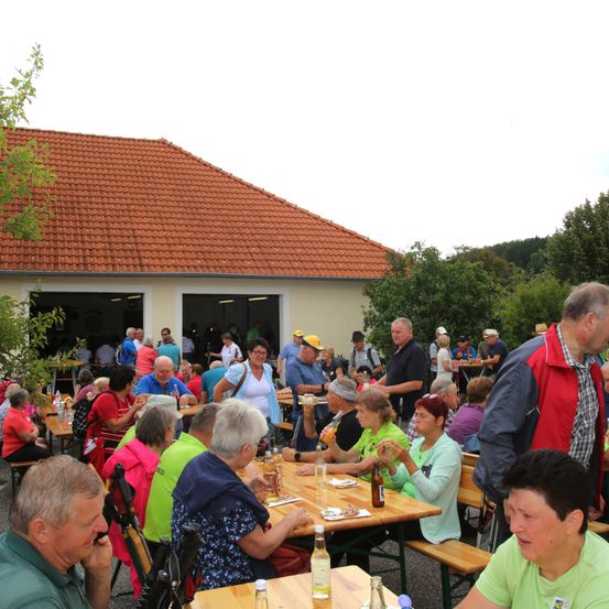 Eine Gruppe von Menschen sitzt zu einer Mahlzeit im Freien an Holztischen unter einem orangefarbenen Dach. Sie essen und trinken und die Szene findet im Freien in der Nähe eines Gebäudes mit offenen Türen statt.