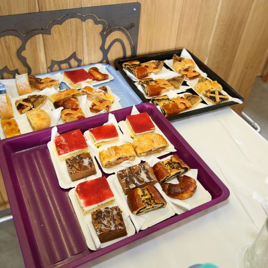 Ein Tisch zeigt mehrere Tabletts mit verschiedenen Gebäcksorten. Das vordere Tablett enthält eine vielfältige Auswahl an Kuchen und Broten, während das hintere Tablett kleinere, mundgerechte Gebäckstücke aufweist.