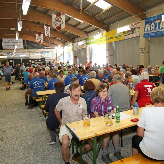 Eine große Menschenmenge sitzt an Tischen in einem Saal. Viele tragen blaue Hemden, und ein Mann mit Schnurrbart sitzt vor einem Tisch mit Getränken.