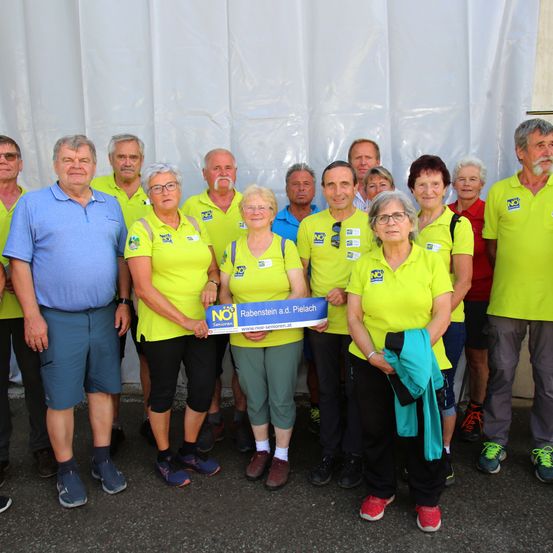 Eine Gruppe von Menschen in gelben Shirts posiert für ein Foto, wobei eine Frau ein blaues Schild mit NO in der Hand hält. Sie stehen vor einem weißen Hintergrund.