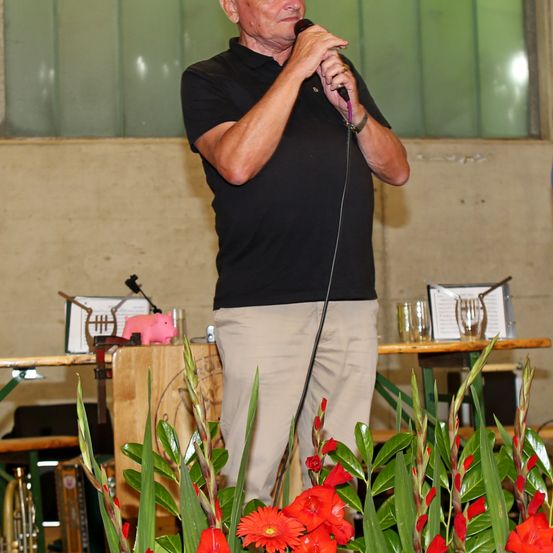 Ein Mann steht und spricht in ein Mikrofon, er trägt ein schwarzes Poloshirt und Khakihosen. Hinter ihm steht ein Tisch mit einem Mikrofon, einem Glas und einer rosafarbenen Sparschweinbank. Der Hintergrund besteht aus einer grünen Wand und einer Holzstruktur.