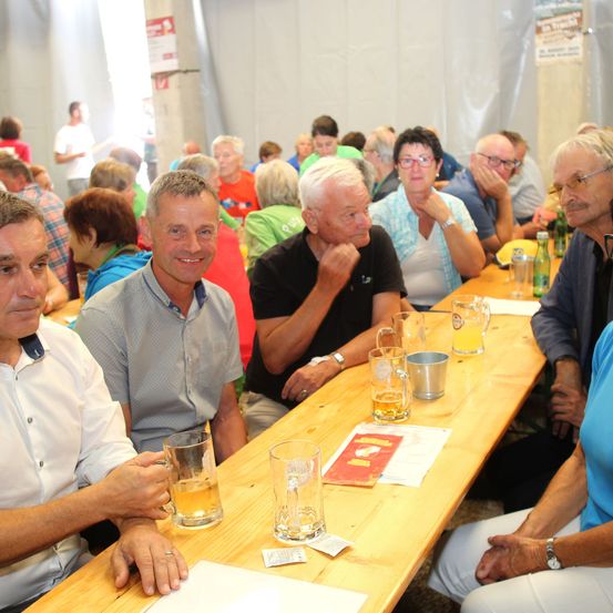 Mehrere Erwachsene sitzen an einem langen Holztisch, einige mit Biergläsern. Die Gruppe scheint zu plaudern und den Moment zu genießen. Eine Person im Vordergrund trägt ein weißes Hemd und hält ein Glas.