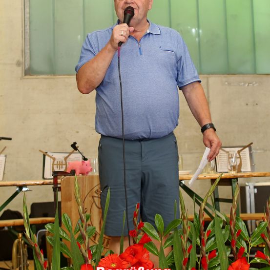 Ein Mann steht vor einem Tisch mit einem Mikrofon und einem Papier in der Hand, trägt ein blaues Poloshirt und graue Shorts. Hinter ihm ist eine grüne Wand und ein Tisch mit einer rosa Schweinefigur und Notenblättern.