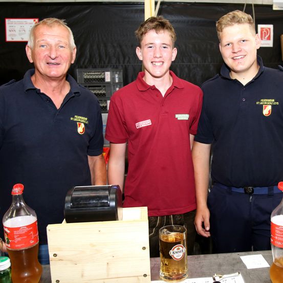 Drei Männer stehen hinter einem Tisch mit Registrierkasse und Getränken, zwei in blauen Poloshirts mit Logos, einer in einem roten Poloshirt.