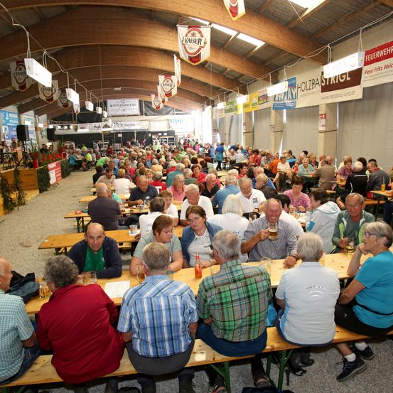 Eine große Menschenmenge sitzt an langen Holztischen in einem großen Zelt. Viele von ihnen halten Gläser mit Bier in den Händen. Werbung und Banner hängen von der Decke.