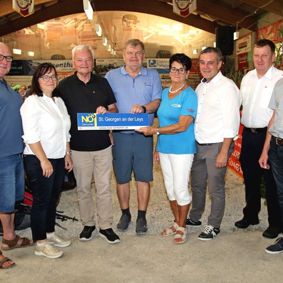 Mehrere Personen posieren für ein Foto mit einem blauen Banner mit der Aufschrift St. Georgen an der Leys. Eine Frau hält einen großen Scheck.