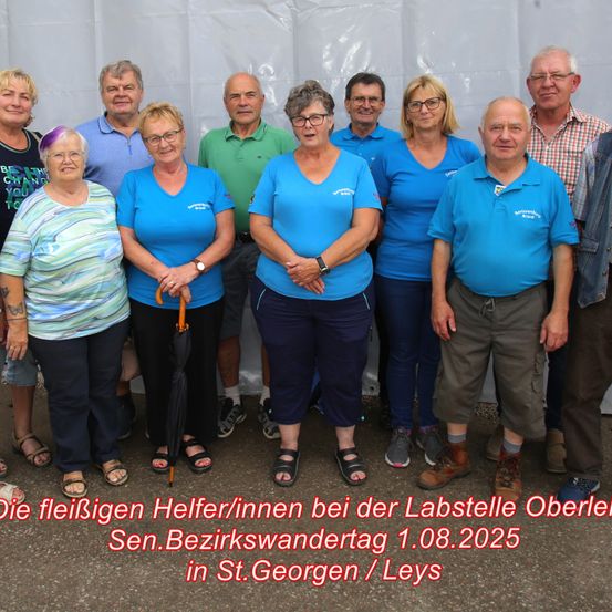 Eine Gruppe von Menschen steht zusammen, lächelt und trägt blaue T-Shirts. Hinter ihnen steht ein weißes Zelt. Der Text lautet: 'Die fleißigen Helfer bei der Labstelle Oberle Sen. Bezirkswandertag 1.08.2025 in St. Georgen / Leys.'