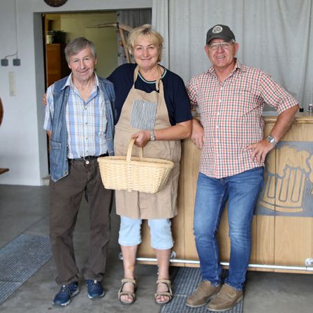 Drei Personen stehen vor einem Tresen. Die Frau in der Mitte hält einen Korb. Beide Männer tragen Freizeitkleidung. Der Tresen hat ein Bierkrug-Design.