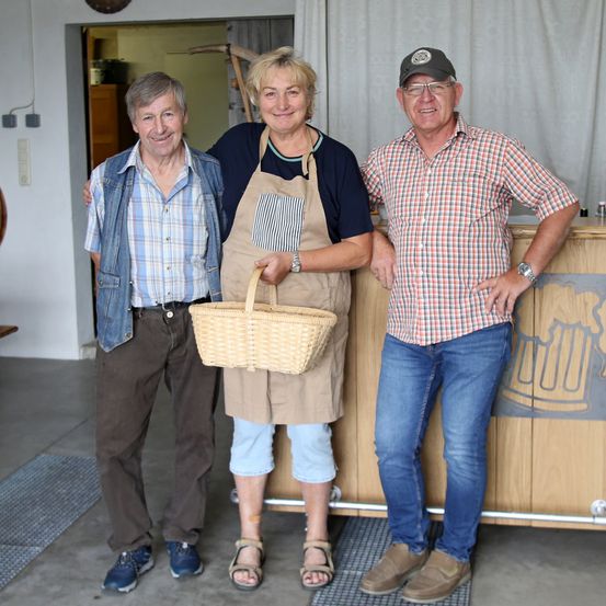 Drei Personen stehen vor einem Tresen. Die Frau in der Mitte hält einen Korb. Beide Männer tragen Freizeitkleidung. Der Tresen hat ein Bierkrug-Design.