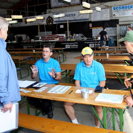 Zwei Männer sitzen an einem Holztisch mit Papieren und einer Karte, einer hält einen Stift. Ein Mann trägt eine gelbe Kappe, der andere ein blaues Hemd. Ein weiterer Mann in einem blauen Hemd hält eine Mappe. Der Hintergrund zeigt eine Küche und andere Tische.
