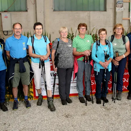 Eine Gruppe von Menschen, gekleidet in Wanderausrüstung, posiert für ein Foto vor einem Gebäude mit einem Schild. Sie lächeln und halten Trekkingstöcke in der Hand.