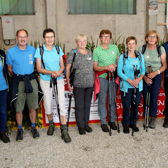 Eine Gruppe von Menschen, gekleidet in Wanderausrüstung, posiert für ein Foto vor einem Gebäude mit einem Schild. Sie lächeln und halten Trekkingstöcke in der Hand.