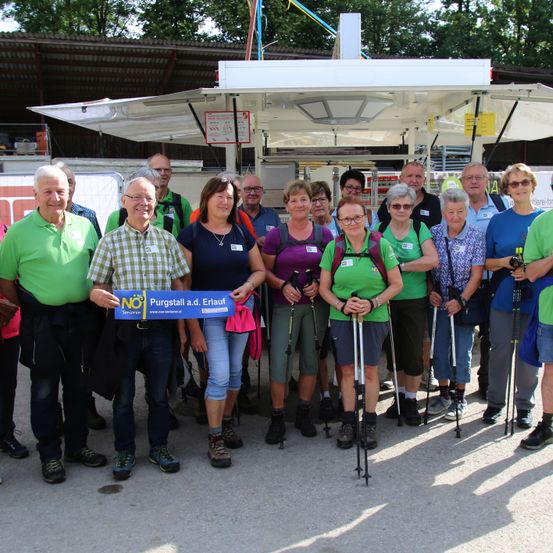 Eine Gruppe von Wanderern steht unter einem Vordach und hält ein Banner. Sie alle lächeln und posieren für ein Foto. Der Hintergrund zeigt eine Struktur mit einem Dach.