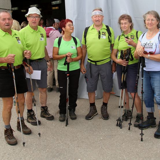 Eine Gruppe von Wanderern in grünen Poloshirts, die Trekkingstöcke halten, posiert für ein Foto. Sie lächeln und schauen nach vorne. Hinter ihnen ist ein weißes Zelt zu sehen, und einige Leute stehen im Hintergrund.