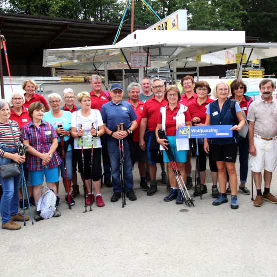 Eine Gruppe von Menschen in roten Hemden und mit Brillen steht vor einem Imbissstand und hält Wanderstöcke. Eine Person hält ein blaues Plakat mit dem Text NEIN! Wolfpassing.