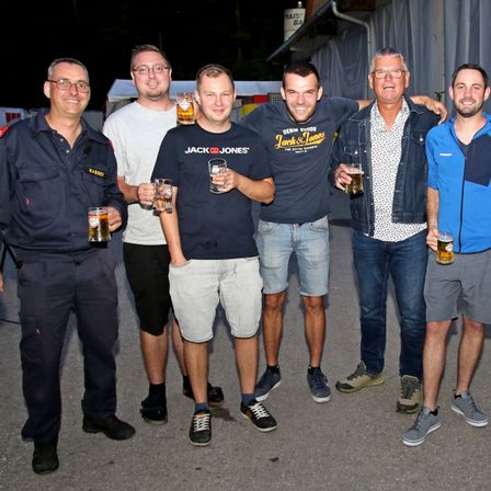 Eine Gruppe von Männern in Freizeitkleidung steht zusammen, lächelt und hält Gläser mit Bier in der Hand. Sie befinden sich wahrscheinlich bei einem geselligen Treffen.