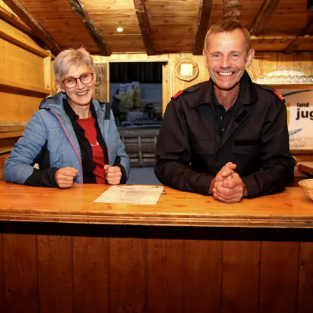 Eine ältere Frau in einer blauen Jacke und einer Brille sitzt hinter einem Schreibtisch, lächelt und posiert mit einem Mann in einer schwarzen Uniform, beide lächeln für ein Foto.