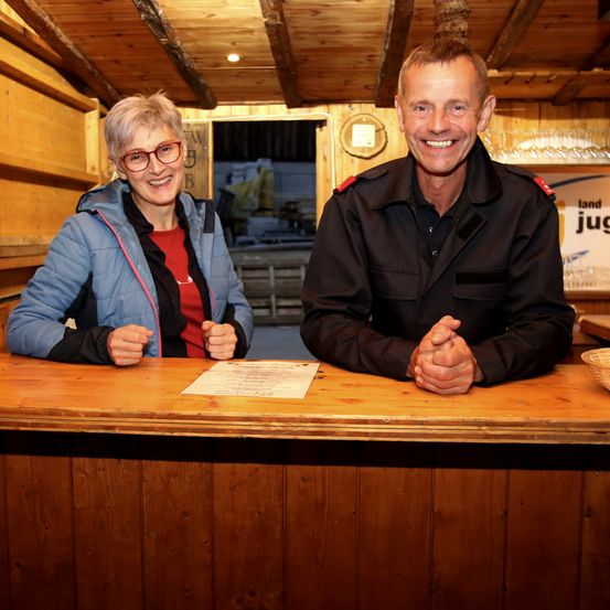 Eine ältere Frau in einer blauen Jacke und einer Brille sitzt hinter einem Schreibtisch, lächelt und posiert mit einem Mann in einer schwarzen Uniform, beide lächeln für ein Foto.