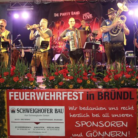 Eine Band spielt auf der Bühne mit Trompete, Akkordeon und Tuba. Die Bühne ist mit roten Blumen dekoriert. Der Hintergrund hat ein Banner mit der Aufschrift 'Die Party Band 100% Live'. Unter der Bühne befindet sich ein Schild mit der Aufschrift 'Feuerwehrfest in Bründl'.