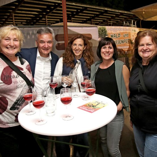 Fünf Erwachsene stehen um einen Tisch mit Gläsern Wein und lächeln für ein Foto. Eine Frau hält eine Flasche Wein. Sie scheinen bei einer Veranstaltung im Freien zu sein.