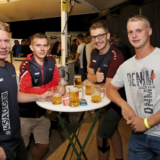 Vier junge Männer bei einem Treffen, zwei tragen eine Brille, einer zeigt den Daumen hoch. Sie halten Biergläser auf einem Tisch, mit einer weiteren Person dahinter.