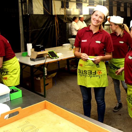 Eine Gruppe von Frauen in Kochuniformen befindet sich in einer Küche. Eine Frau lächelt und hält ein Stück Papier in der Hand. Hinter ihr tragen zwei weitere Frauen Schürzen. Vor ihnen steht ein Tisch mit verschiedenen Gegenständen und einem Telefon.