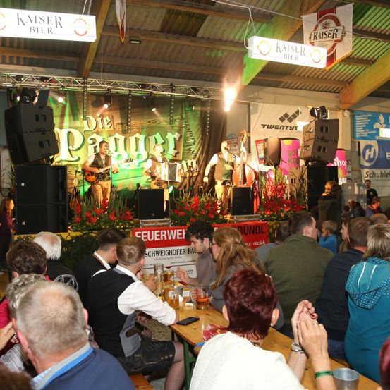 Eine Band spielt auf einer Bühne bei einer Indoor-Veranstaltung. Sie haben Lautsprecher, ein Banner und eine grüne Leinwand mit Text. Das Publikum sitzt und schaut zu.