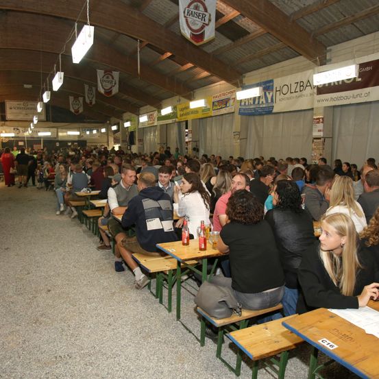 Eine große Gruppe von Menschen sitzt an Tischen in einer Halle, wahrscheinlich bei einem Festival. Die Tische sind aus Holz und die Leute unterhalten sich. Mehrere Banner hängen an den Wänden.