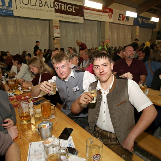 Zwei Männer in traditioneller bayerischer Kleidung sitzen an einem Tisch in einem überfüllten Biergarten und halten Biergläser in der Hand. Der Tisch ist mit verschiedenen Gläsern und einem Handy bestückt. Der Raum ist mit anderen Menschen gefüllt und die Wände haben Banner und Schilder.