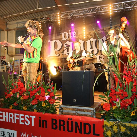 Eine Band spielt auf der Bühne mit rotem Hintergrund. Der Leadsänger trägt ein farbenfrohes Shirt, während zwei Bandmitglieder Gitarren spielen. Ein großer Lautsprecher steht vor ihnen. Bühnenlichter sind sichtbar, und es gibt ein Schild, das 'Festival in Brundl' sagt.