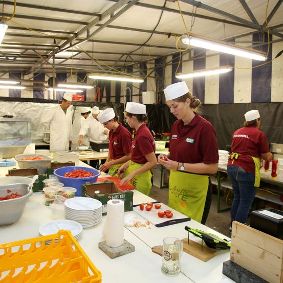 Eine Gruppe von Köchen in einer Küche bereitet Essen zu, wobei eine Frau in einem roten Hemd und gelber Schürze eine Tomate untersucht, während andere hinter ihr arbeiten, und ein Tisch voller Teller und Zutaten.