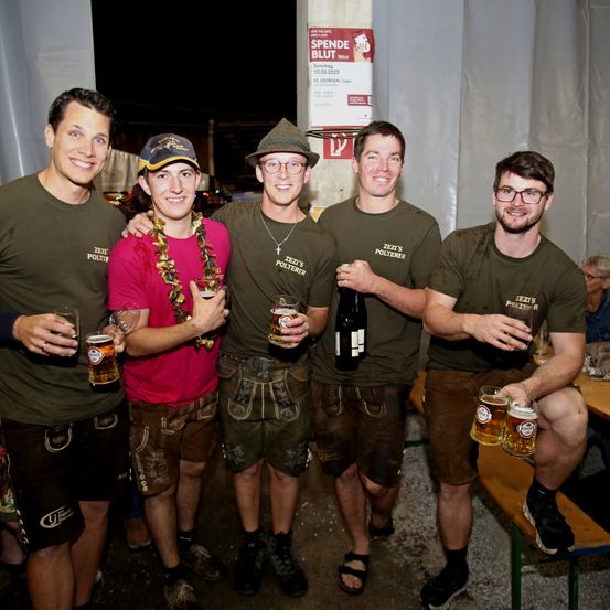 Fünf Männer in grünen Uniformen und braunen Shorts posieren nachts für ein Foto. Sie halten Gläser mit Bier und eine Flasche Wein. Ein Schild an der Wand steht 'Spende Blut'.