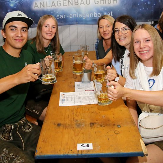 Eine Gruppe junger Leute sitzt um einen Holztisch mit Bierkrügen, lächelt und posiert für ein Foto. Ein Schild auf dem Tisch steht 'FE'.